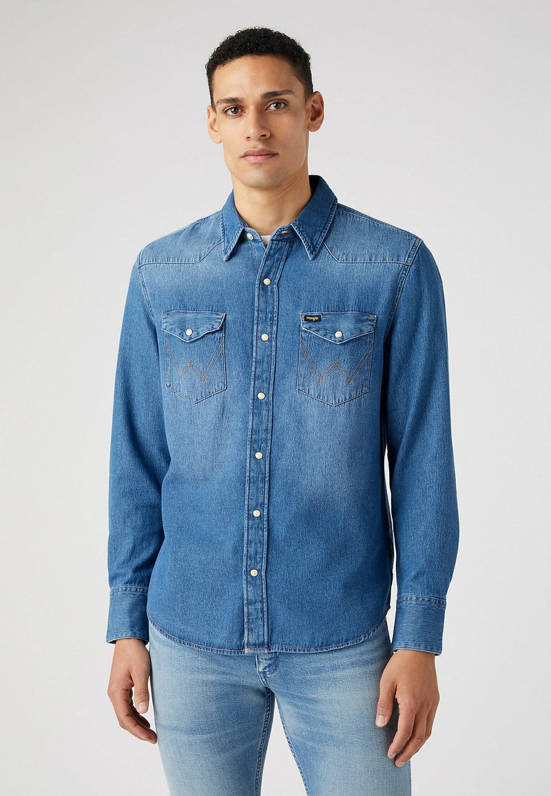Camisa de mezclilla en azul medio con cierre de botones, cuello extendido, dos bolsillos delanteros y detalles de costura en contraste. Mangas largas, corte relajado.