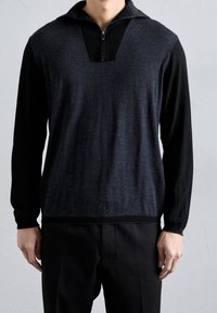 Pull-over gris foncé à fermeture éclair mi-hauteur avec manches longues noires. Présente un tricot texturé, un col en V et une silhouette ajustée.