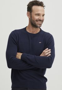 Mornarsko moder pulover s krožnim izrezom, narejen iz pletenega blaga, ki ima na prsih majhen logotip v rdeči, beli in modri barvi, z rebrastimi manšetami in robom.