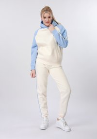 Jeune femme portant un sweat à capuche crème et bleu clair avec un jogging assorti et des baskets blanches, debout devant un fond uni clair.