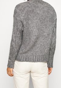 Osoba oblečená v šedém pleteném svetru s kabelkovými vzory na rukávech a ribovými manžetami, v kombinaci se světle béžovými kalhotami, pohledem zezadu.