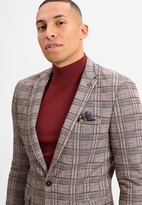 Kariertes Blazer in Beige, Rot und Marineblau, mit einer Zweiknopfverschluss und einem Einstecktuch mit buntem Muster. getragen über einem bordeauxfarbenen Rollkragenpullover.