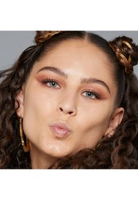 In primo piano, una persona con capelli ricci, che indossa orecchini a cerchio dorati, con labbra lucide e un leggero ombretto marrone, che fa un'espressione di bacio.