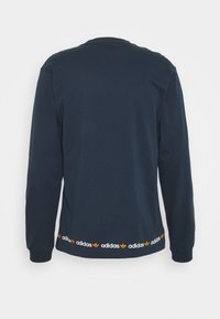 Námořnické tričko s dlouhým rukávem z bavlny s kulatým výstřihem. Na spodním lemu má pruh s logem Adidas v oranžové a žluté barvě. Jednoduchá, hladká textura.