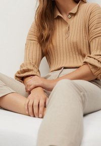 Polo beige côtelé avec manches retroussées et détails à boutons, associé à un pantalon beige clair. Texture lisse, coupe ajustée.