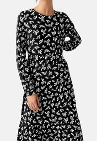 Robe noire à manches longues, motif floral blanc, col rond, taille froncée et ourlet à volants. Fabriquée en matériau léger et doux.