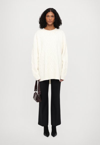 Pull crème en maille torsadée surdimensionné, pantalon large noir, escarpins noirs à bout pointu et un petit sac à main bordeaux brillant.