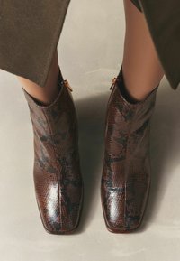 Brūni un melni čūsku raksta potītes zābaki ar kvadrātveida purngalu un zelta rāvējslēdzēja akcentiem sānos. Gluda ādas textura, elegants dizains.