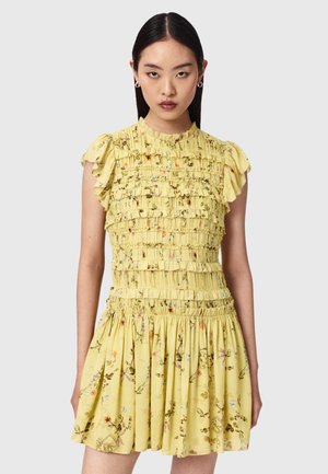 Jeune femme aux cheveux noirs et lisses portant une robe courte jaune à motif floral avec des détails à volants et des manches courtes bouffantes, sur un fond gris.