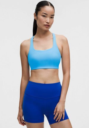 Mujer joven con cabello largo y oscuro recogido en una coleta, vistiendo un sujetador deportivo azul claro y pantalones cortos deportivos azul real de cintura alta, de pie frente a un fondo liso.