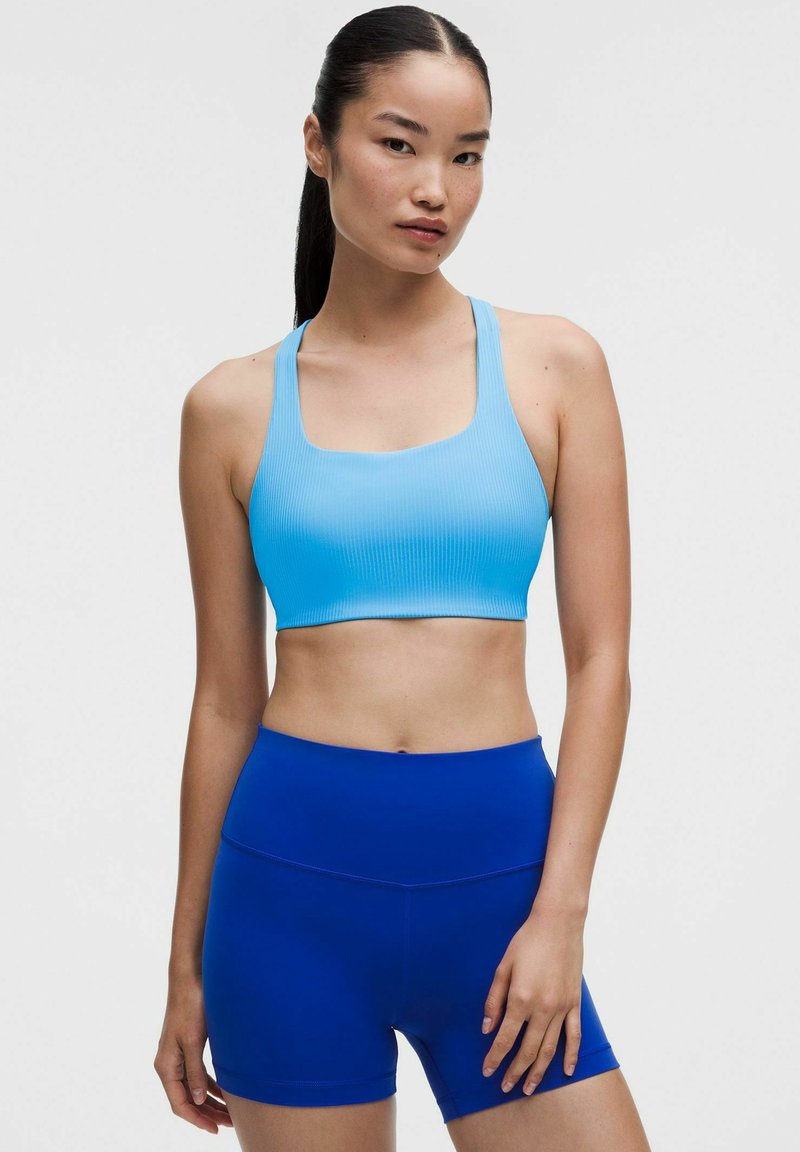 Young woman with long dark hair in a ponytail wearing a light blue sports bra and royal blue high-waisted athletic shorts standing against a plain background.