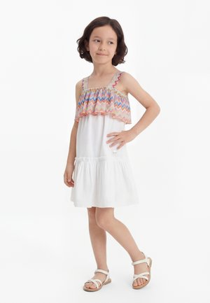 Fille debout, main sur la hanche, portant une robe blanche avec un volant zigzag coloré et des sandales blanches sur fond blanc.