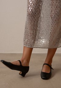 Svarta läder Mary Jane-skor med rund tå och ett rem över vristen. Låg klack och slät yta. Bärs med en klänning i silverfärgade paljetter.