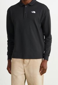 Mörk polotröja med långa ärmar i svart med en mönstrad textur, med en vit logotyp på vänster bröst, kombinerat med ljusa beige byxor.