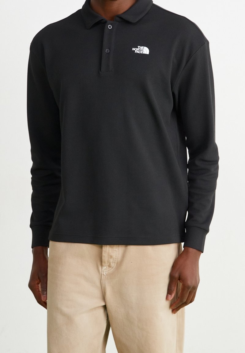 Mörk polotröja med långa ärmar i svart med en mönstrad textur, med en vit logotyp på vänster bröst, kombinerat med ljusa beige byxor.