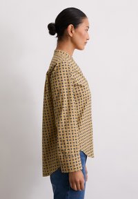 Langsleeve shirt in mosterdgeel met een marineblauw geometrisch patroon, voorzien van een kraag en een ontspannen pasvorm, gedragen met blauwe jeans.