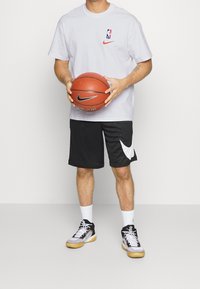 Camiseta blanca de manga corta con los logotipos de la NBA y Nike, pantalones cortos de baloncesto negros con el swoosh blanco de Nike, y zapatillas de baloncesto de caña alta en negro/gris.