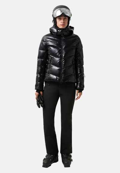 Zwarte donzen jas met glanzende afwerking, ritssluiting en capuchon; gecombineerd met zwarte skibroek en skischoenen; gecompleteerd met handschoenen en skibril.