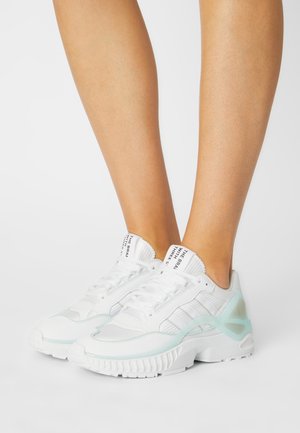 Une personne portant des baskets de sport blanches et bleu clair, debout sur un fond blanc uni.