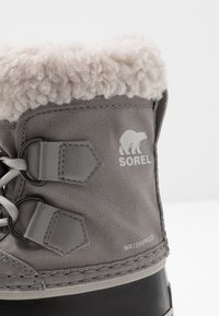 Bota cinza impermeável com forro de pele sintética, parte superior em tecido texturizado e sola de borracha preta. Apresenta detalhes em hardware hexagonal e logotipo.