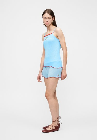 Γυναίκα που φοράει ανοιχτό μπλε καμησόλ και σορτς με κόκκινο περίγραμμα και μικρά κεντητά σχέδια, συνδυασμένα με καφέ δερμάτινα παπούτσια με κορδόνια.