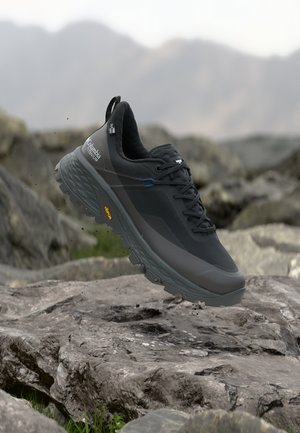 Črna planinska obutev Columbia Titanium z Vibram podplatom lebdi nad skalnatim terenom, v ozadju pa so gore.