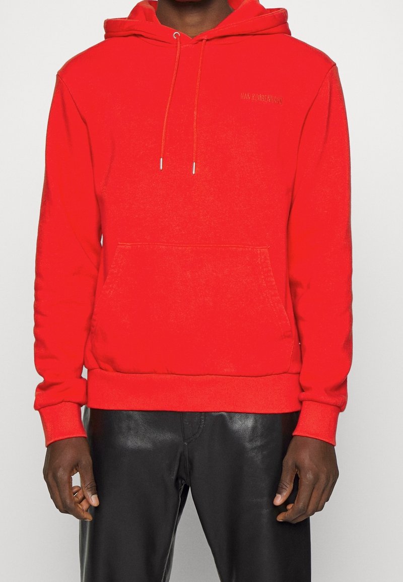 Homme portant un sweat à capuche rouge vif avec poche avant et pantalon en cuir noir sur fond uni.