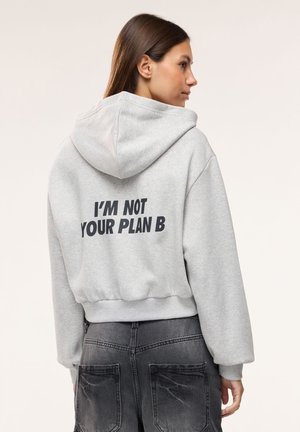 Młoda kobieta z długimi brązowymi włosami, ubrana w jasnoszarą bluzę z kapturem z napisem "I'M NOT YOUR PLAN B", stoi na tle jednolitego tła.