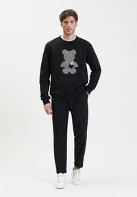 Zwarte sweatshirt met een grote berenprint in contrasterend materiaal, gecombineerd met zwarte broek en witte sneakers. Casual, relaxte pasvorm.