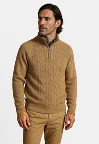 Pull en tricot beige avec un col montant et une fermeture éclair ; présente un motif en maille torsadée ainsi que des bords côtelés aux poignets et à l'ourlet. Porté avec une chemise à col.