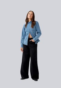 Light blue denim shirt with buttons, collar, and open side detail, paired with high-waisted, wide black pants. Smooth texture, casual fit.