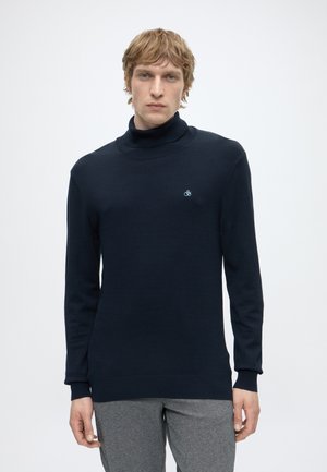 Young man with blonde hair wearing a fitted navy turtleneck sweater and gray pants, standing against a plain light background.