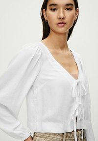 Hvid cropped bluse lavet af linned med dyb V-hals, puffede ærmer og frontbinding. Glat tekstur med minimale designaccenter.
