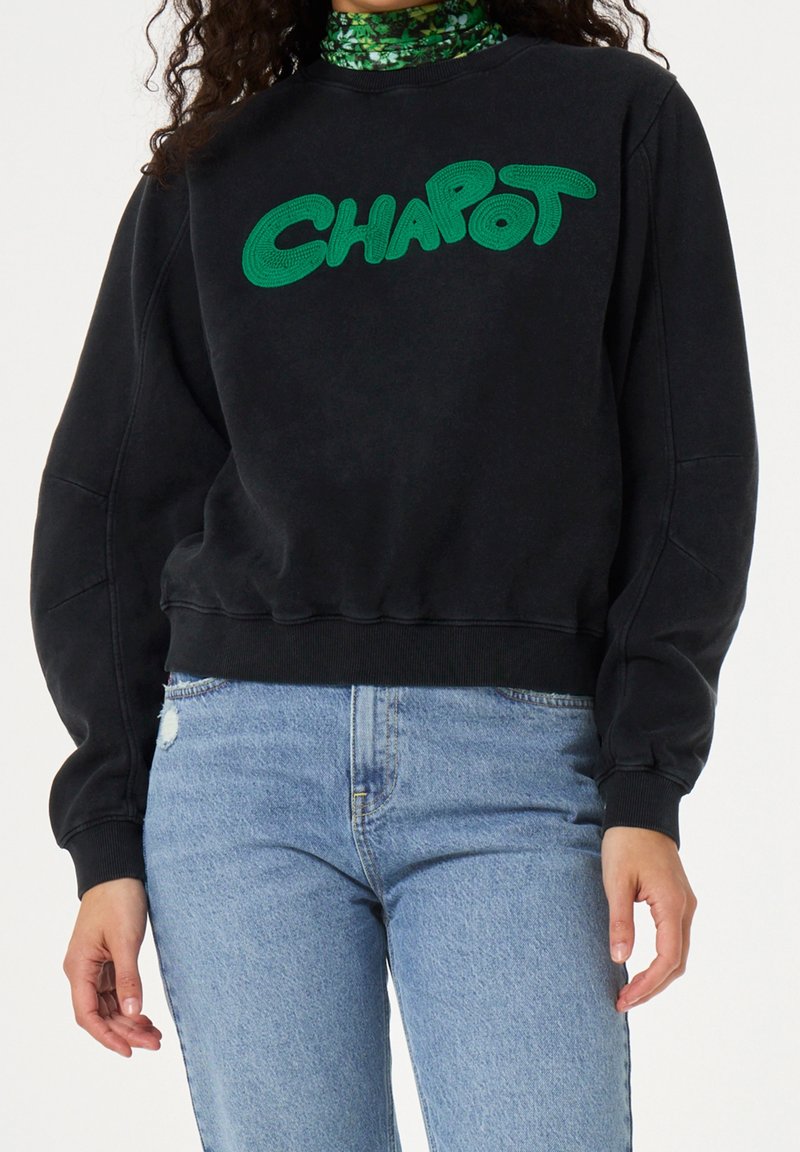 Persoon draagt een zwarte trui met groene "CHAPOT" tekst en lichtblauwe spijkerbroek, tegen een effen witte achtergrond.