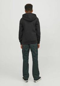 Felpa con cappuccio nera e pantaloni cargo verde scuro con cuciture bianche. Sneakers nere con suole bianche e lacci neri. Vista da dietro.
