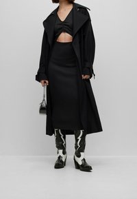 Trench-coat noir porté sur un haut noir ajusté et une jupe, assorti à des bottes montantes noires et blanches ornées d'éclairs. Petit sac noir avec chaîne.