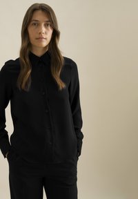 Zwarte button-up shirt met lange mouwen, gladde textuur, spitse kraag en een rechte pasvorm. Gecombineerd met zwarte broeken, wat een monochrome uitstraling creëert.