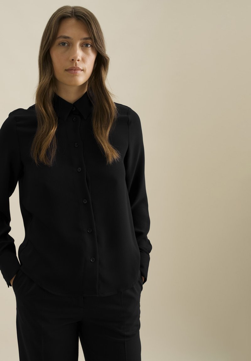 Zwarte button-up shirt met lange mouwen, gladde textuur, spitse kraag en een rechte pasvorm. Gecombineerd met zwarte broeken, wat een monochrome uitstraling creëert.