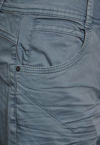 Niebieskie spodnie bawełniane z dodatkiem innych włókien o gładkiej strukturze, z ukośnymi kieszeniami, widocznym szwem i delikatnym połyskiem. Idealne do codziennych stylizacji.