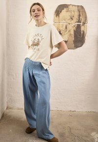 Femme debout à l'intérieur, portant un t-shirt crème "Coastal Breeze", un pantalon bleu ample et des chaussures marron, avec une œuvre murale abstraite derrière elle.