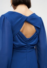 Γυναίκα που φοράει μπλε φόρεμα με μακριά διαφανή μανίκια και σπειροειδές ανοιχτό πίσω μέρος, αποκαλύπτοντας δέρμα ανάμεσα στις πτυχές του υφάσματος.