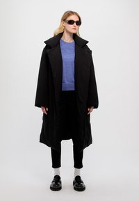Svart oversized kappa med bred krage, blå skjorta under, svarta byxor, svarta skor och svarta solglasögon. Slät tyg, avspänd passform.