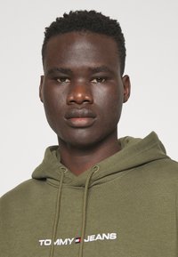 Sudadera con capucha de color verde oliva con cordones, que presenta el logo blanco "TOMMY JEANS" y acentos en rojo, blanco y azul. Textura de tela suave.