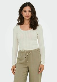 Langarmshirt aus off-weißem Rippstoff mit einem gewellten Ausschnitt, kombiniert mit hellbeigen Hose mit Kordelzug.