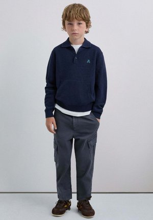 Sudadera azul marino con cuello y bolsillo frontal, de punto texturizado, combinada con pantalones cargo grises y zapatos marrón oscuro con acentos amarillos.