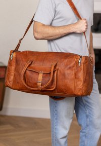 Brun læderet duffel-taske med dobbelte håndtag og en aftagelig skulderrem. Har en forlomme, sølvfarvede lynlåse og en tekstureret overflade.