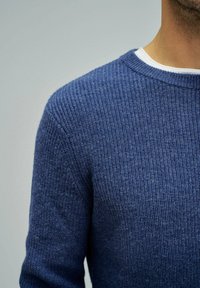 Maglione lavorato a maglia blu scuro con texture a costine, colletto rotondo e maniche lunghe, indossato sopra una camicia bianca.