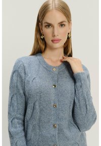 Niebieski sweter robiony na drutach z miękkiej wełny, z okrągłym dekoltem, zapięciem na guziki oraz teksturowanymi wzorami na rękawach.