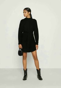 Čierne pletené šaty s dlhými rukávmi, s úzkym pásom a okrúhlym výstrihom. Nosené s členkovými čižmami a malou kabelkou. Hladká textúra.