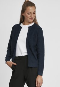 Cardigan en tricot bleu marine avec un design en relief à pompons, encolure ronde et devant ouvert. Assorti à une chemise blanche et un pantalon noir.